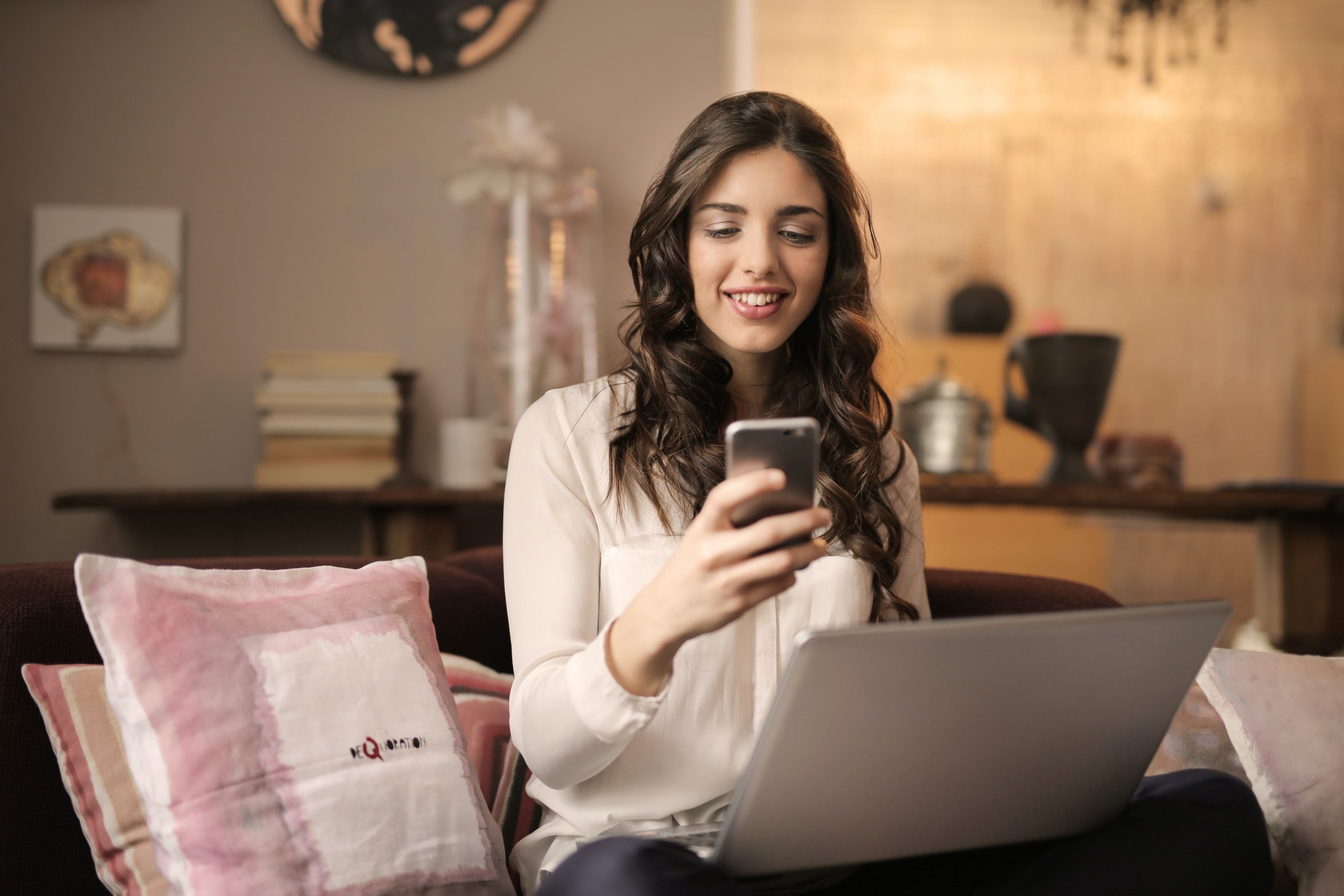 woman smiling while sitting on the sofa, holding a mobile phone with a laptop on the lap
